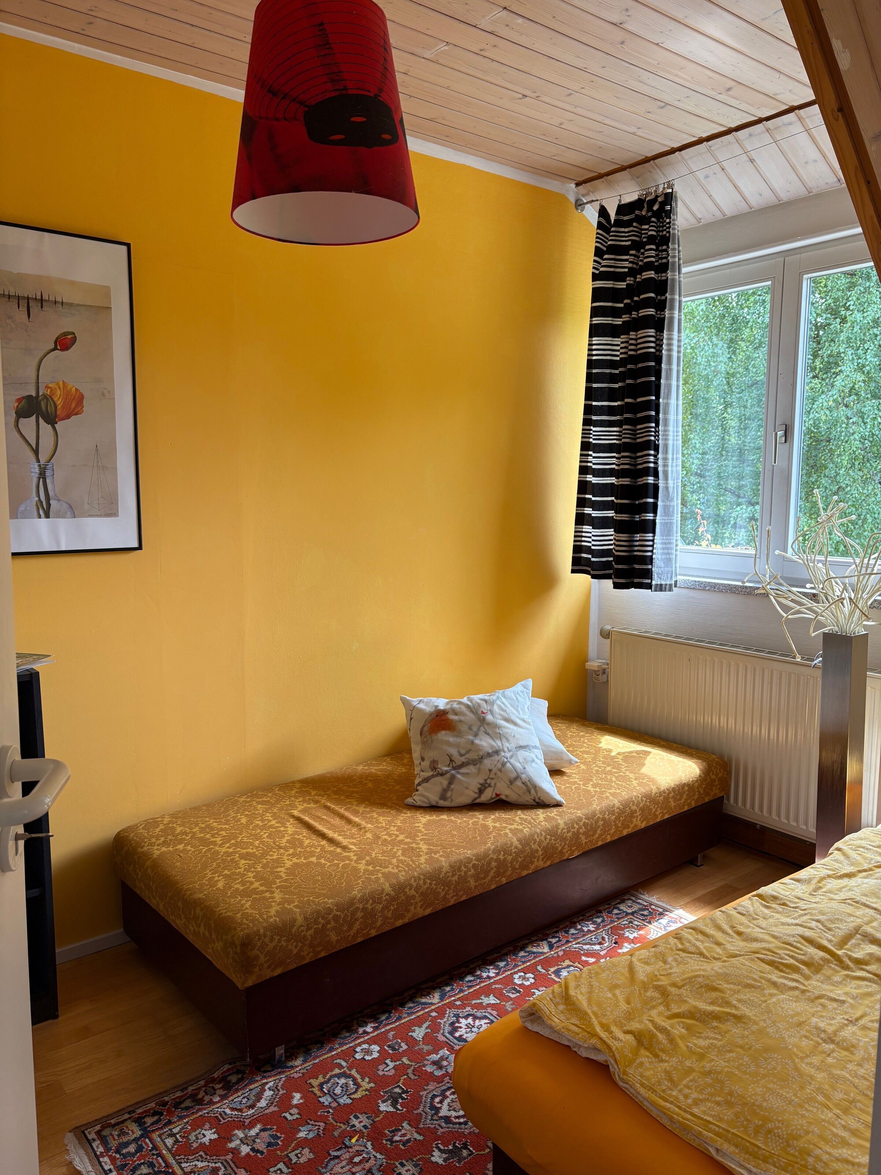 Een kamer met een bed, een raam met gordijnen, een radiator en een schilderij aan de muur.