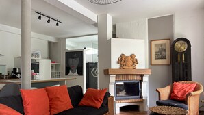 Un salon avec un canapé noir, des coussins rouges, une cheminée, une horloge et un fauteuil en osier.