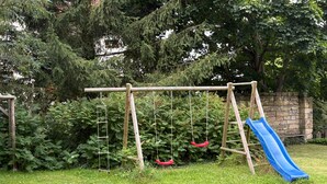 Un ensemble de jeux d’extérieur en bois, avec un toboggan bleu, installé dans une cour arrière verdoyante, agrémentée d’arbres et d’un mur en pierre.