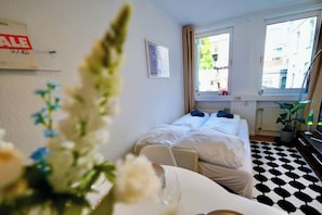 A hotel room with a bed, a window, a radiator, a potted plant, and a table with a glass.
