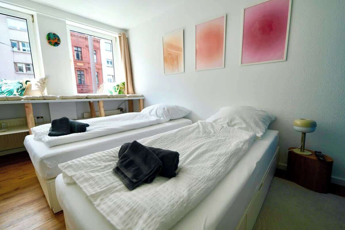 Ein Hotelzimmer mit zwei Betten, einem Fenster mit Blick auf Gebäude und einem Nachttisch mit einer Lampe.
