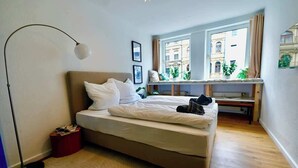 A bedroom with a bed, a bedside table, a lamp, a window with a view of buildings, and a small plant on the windowsill. - Modernes Apartment direkt im Szeneviertel Kölns (Köln)
