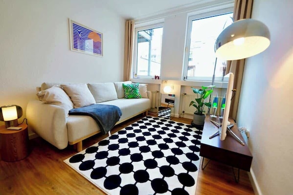 A modern living room with a sofa, a patterned rug, a desk with a lamp, and a window with curtains.