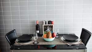 Une table à manger avec des chaises noires, un saladier de fruits, une bouteille de vin et une petite étagère contenant des ustensiles de cuisine.
