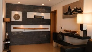 Un hall d’entrée moderne avec un mur en pierre sombre, un éclairage encastré, un banc en bois, un lampadaire et une œuvre d’art encadrée.