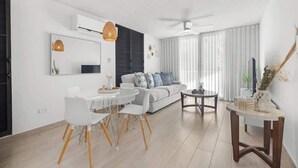 Ein modernes Wohnzimmer mit einem weißen Sofa, einem Esstisch mit Stühlen, einem Deckenventilator und einem großen Fenster mit Vorhängen.
