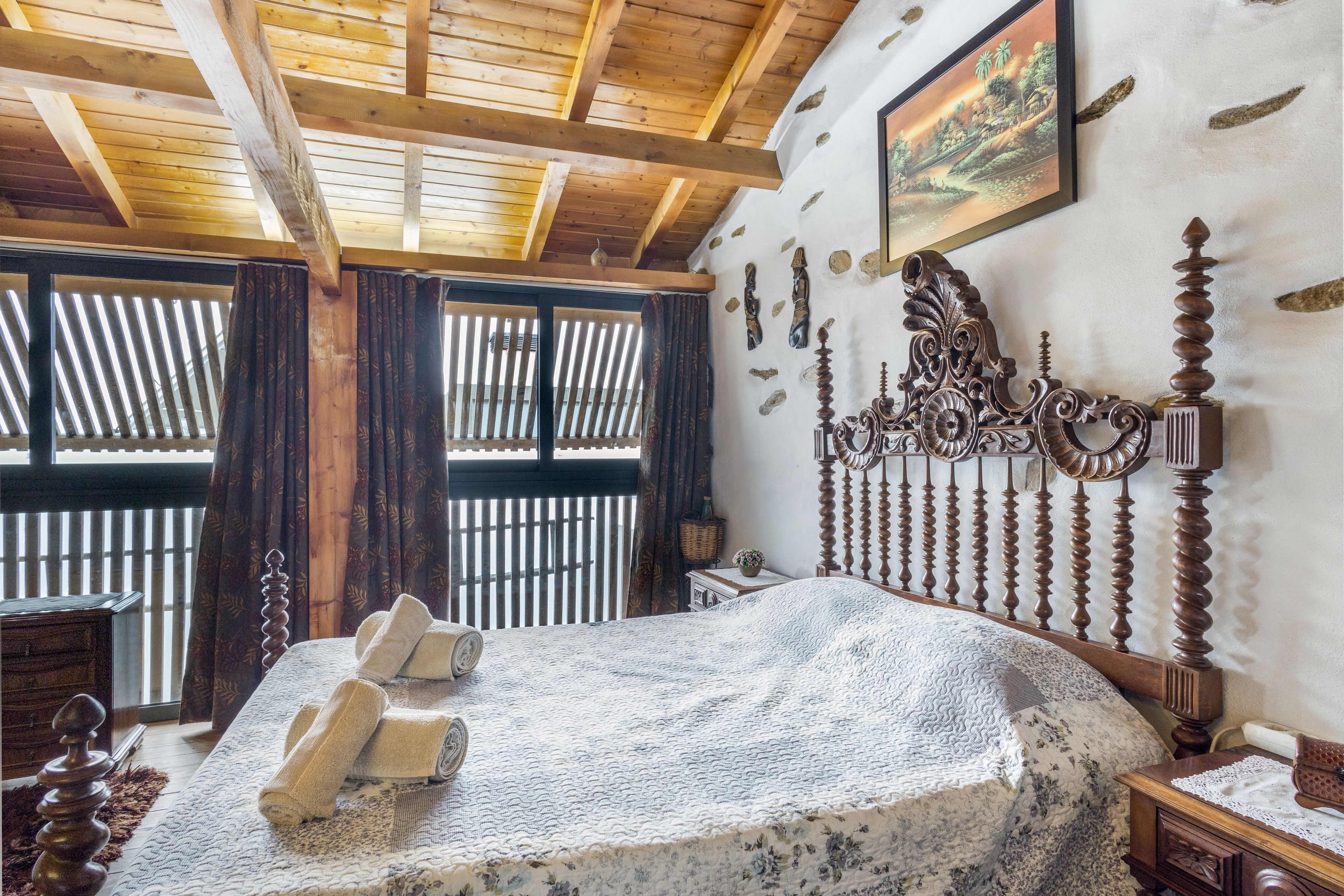 A bedroom with a wooden ceiling, a bed with a decorative headboard, a window with curtains, and a wall-mounted picture.