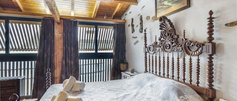 A bedroom with a wooden ceiling, a bed with a decorative headboard, a window with curtains, and a wall-mounted picture.