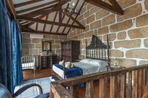 Een slaapkamer met een stenen muur, een houten plafond, een bed met hoofdbord, een dressoir, een spiegel en een raam met gordijnen.
