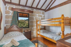 Une chambre avec un mur en pierre, des lits superposés en bois, un lit simple, une table de chevet, un miroir et une fenêtre donnant sur un paysage verdoyant.