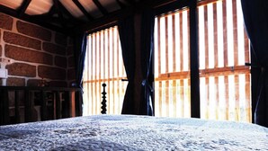 A room with a stone wall, a bed with a patterned bedspread, and a window with vertical blinds.