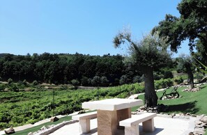 Um banco de pedra com uma mesa num jardim ajardinado, com árvores e uma cerca ao fundo.