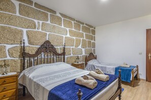 A bedroom with a stone wall, two beds with wooden headboards, a nightstand, a dresser, and a door. - Casa Do Vale JC - Sobreiro (Vila Nune)