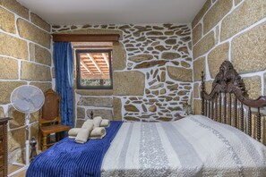 A room with stone walls, a bed with a patterned bedspread, a wooden headboard, a window with a wooden shutter, and a standing fan.