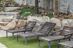Drie houten lounge stoelen met metalen frames, geplaatst op een grasveld met een stenen muur en trappen op de achtergrond.