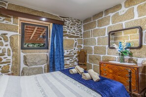 Une pièce aux murs de pierre, avec un lit en bois, une commode, un miroir et une fenêtre donnant sur un paysage verdoyant.