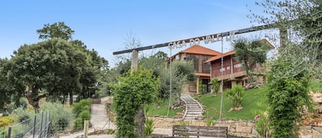 A house with a stone staircase leading to a red-roofed building, surrounded by a well-maintained garden with various plants and a swing set.