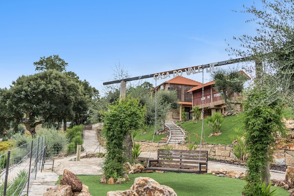 A house with a stone staircase leading to a red-roofed building, surrounded by a well-maintained garden with various plants and a swing set.