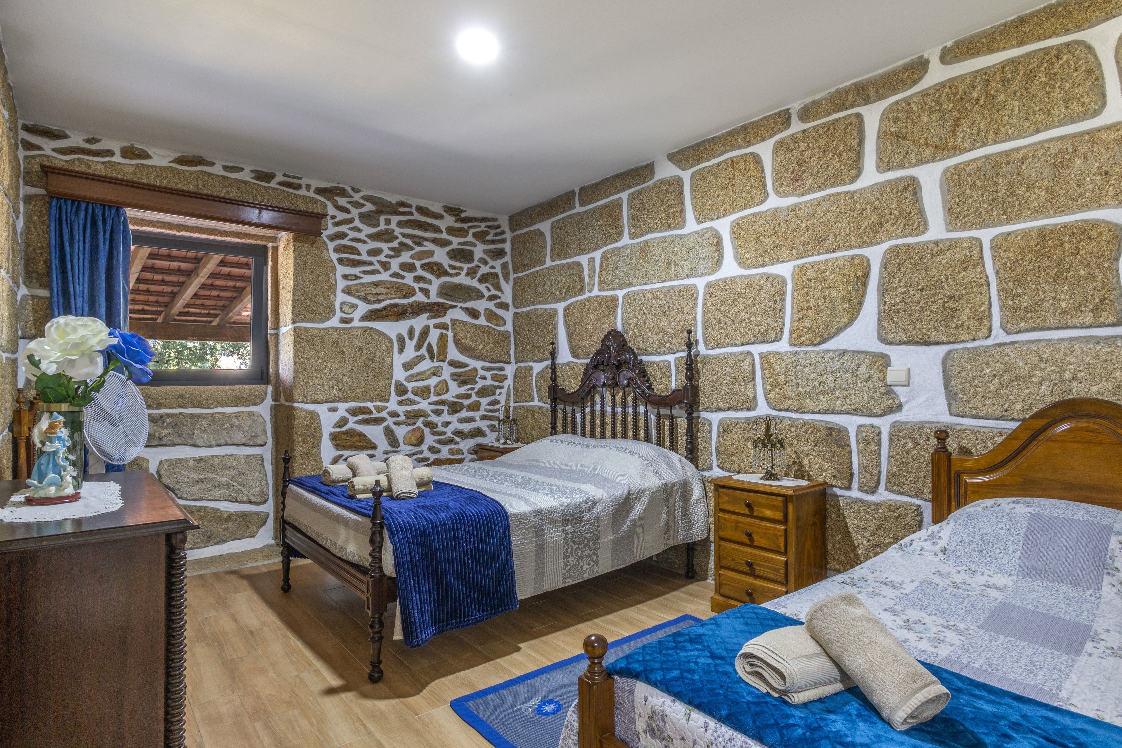 A bedroom with a stone wall, two beds, a wooden dresser, and a window with curtains.