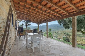 A covered patio with stone flooring, a table with chairs, and a view of a hilly landscape.