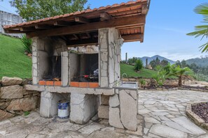 A stone-built outdoor barbecue area with a metal grill, a stone barrel, and a stone wall. - Casa Do Vale JC - Sobreiro (Vila Nune)