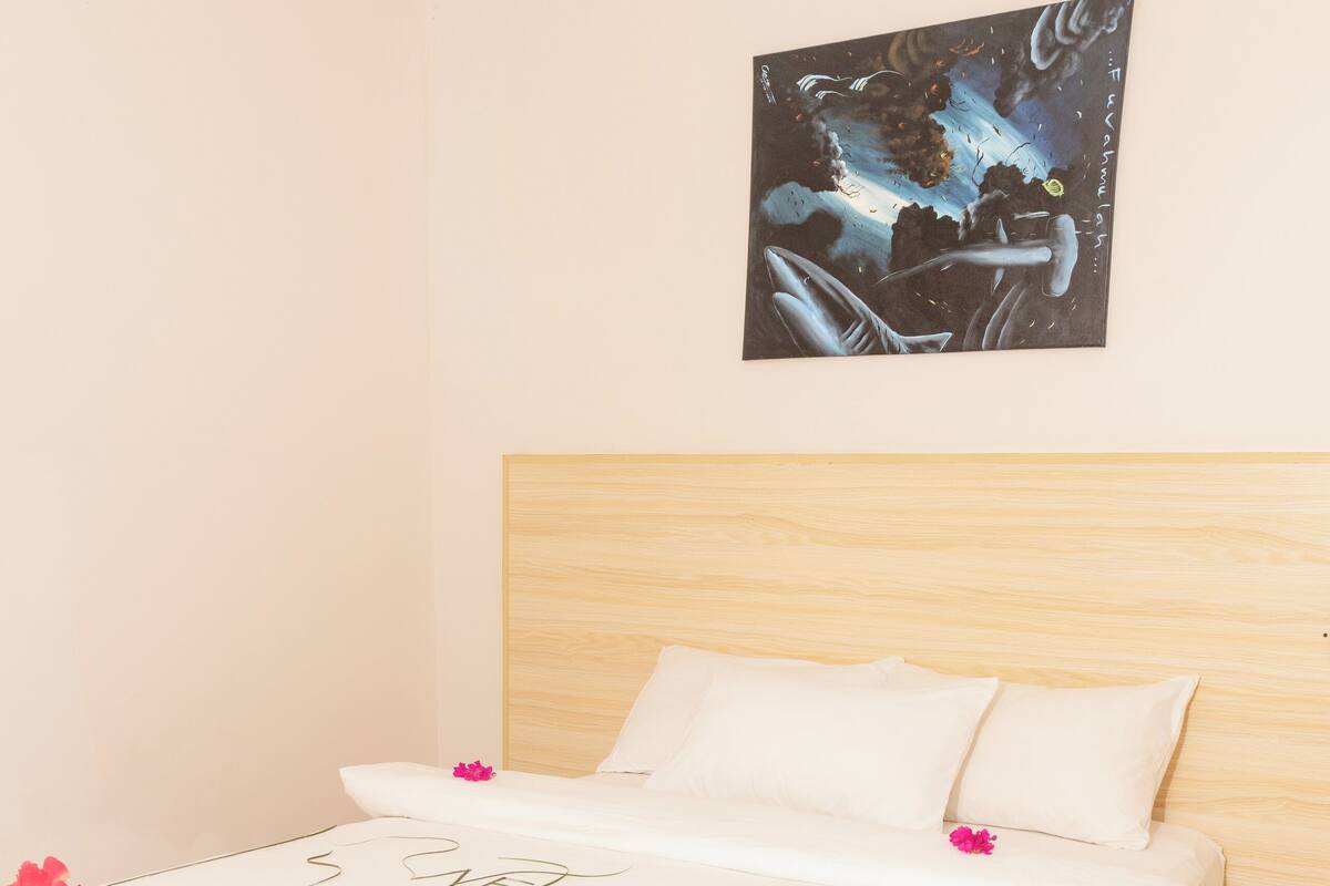 A neatly made bed with white linens and pink flowers, a wooden headboard, and a painting on the wall.