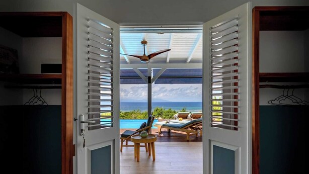 An open door leading to a balcony with a view of the ocean, a ceiling fan, and a table with chairs.