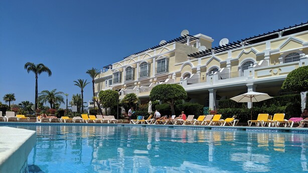 Une piscine avec des chaises longues et, au fond, un grand bâtiment doté de balcons.
