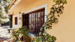 A yellow building with a large window, surrounded by lush greenery and a small patio area. - Los Almendros. (Valsequillo)