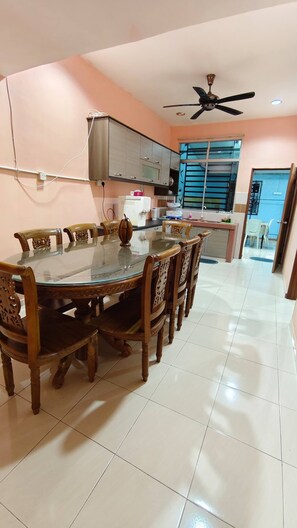 A dining area with a round glass table surrounded by wooden chairs, a ceiling fan, and a kitchen in the background. - Homestay Murah Cheap and Very Satisfying (Kuantan)
