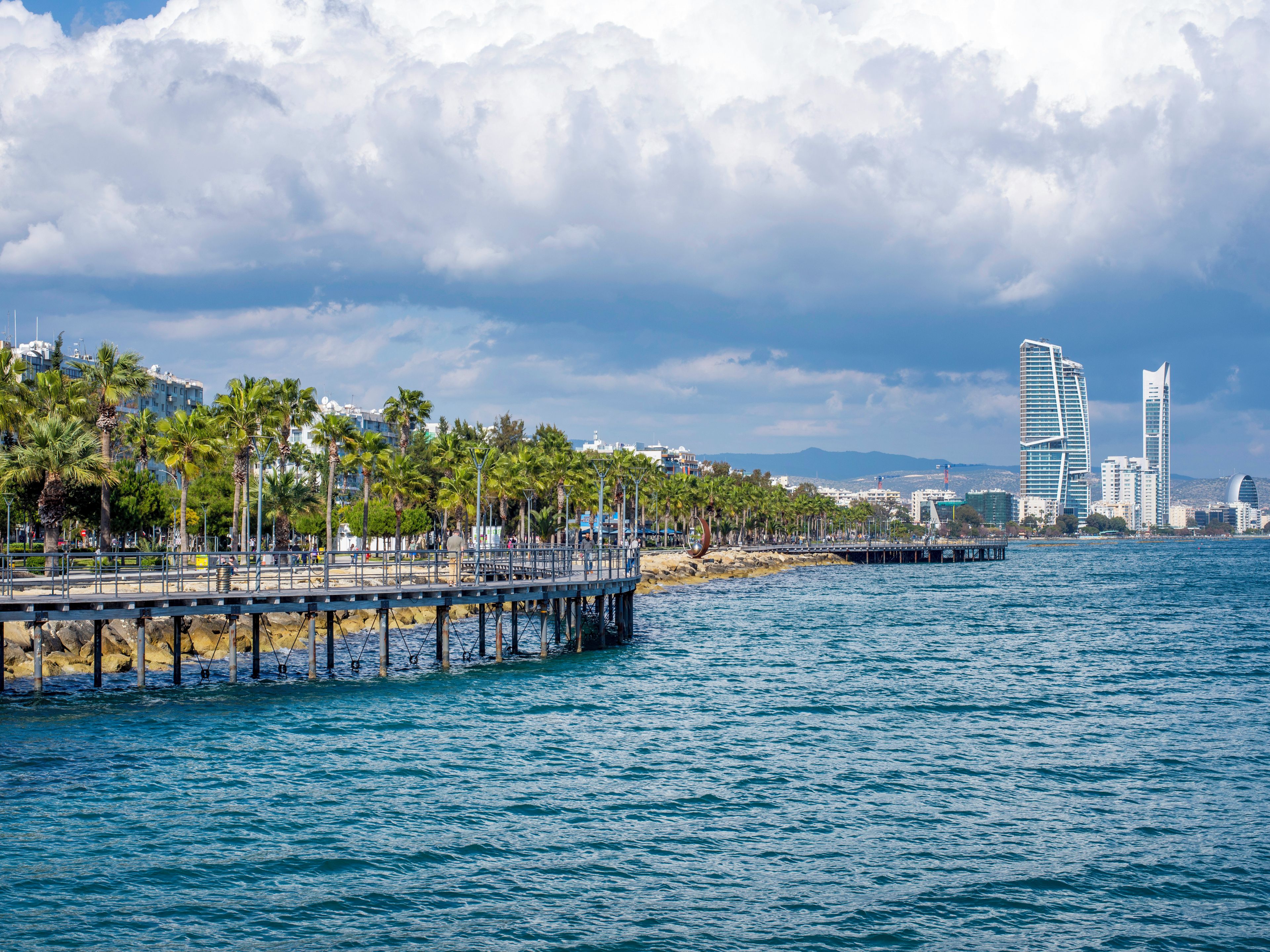 A coastal cityscape with a pier extending into the sea, lined with palm trees and modern buildings in the background.