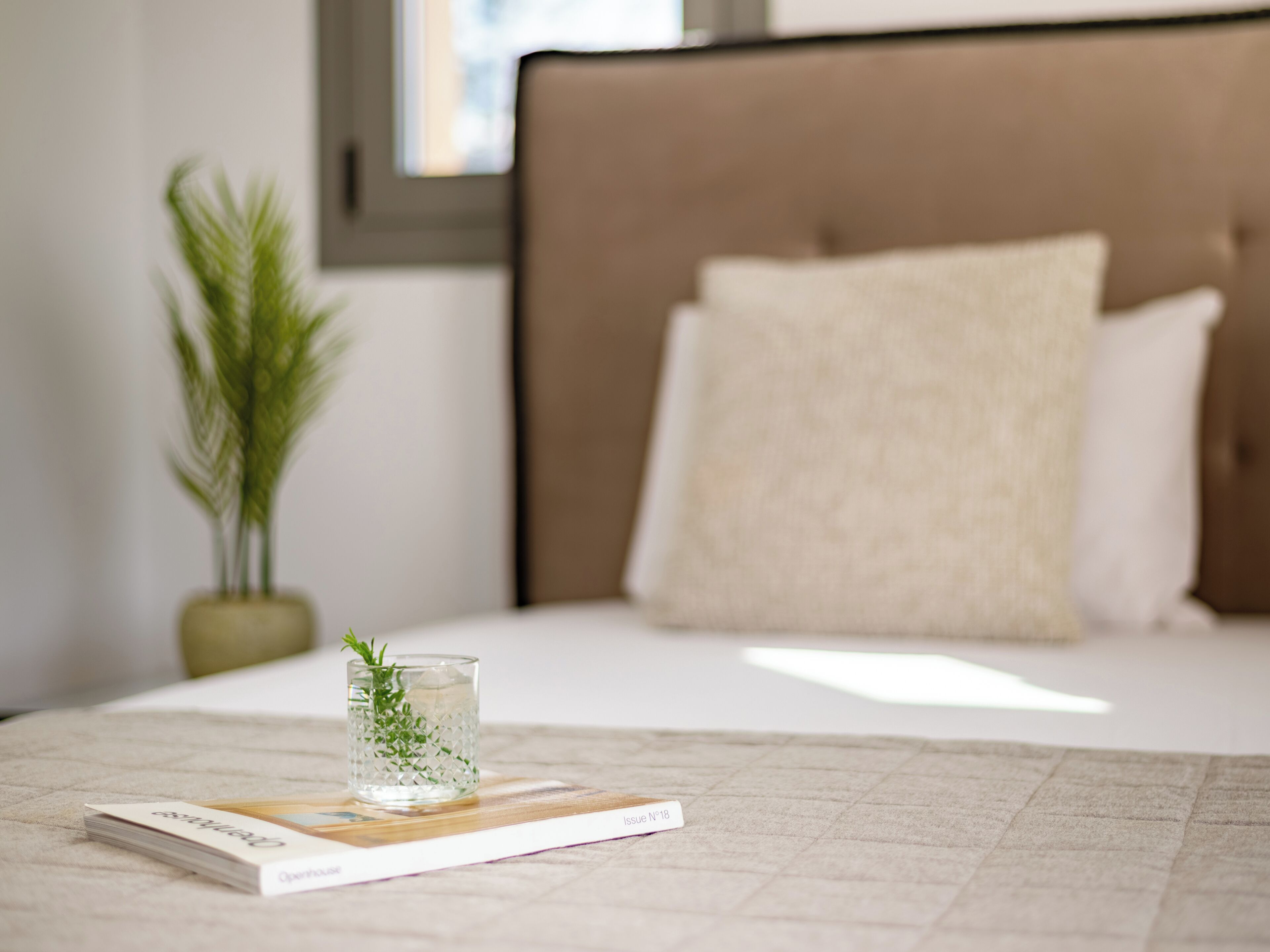 A neatly made bed with a beige pillow and a glass of water on a magazine.