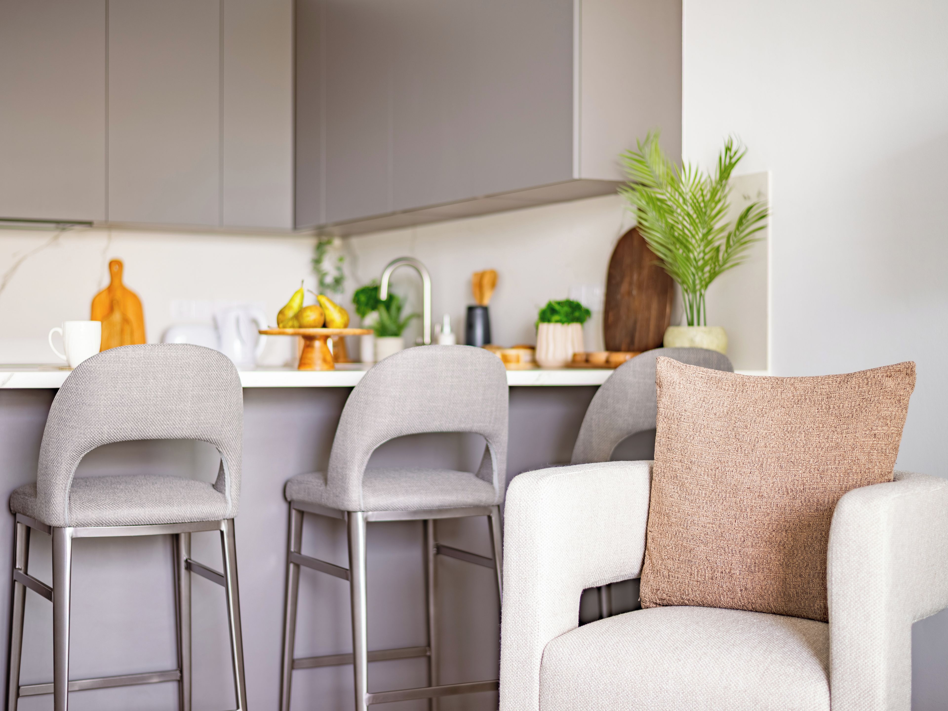 A modern kitchen with a white countertop, two grey bar stools, and a white armchair with a textured pillow.