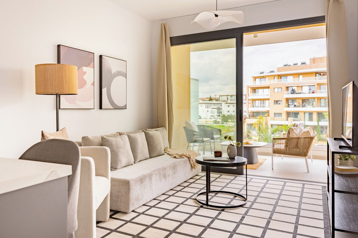 A modern hotel room with a sofa, a small table, a TV, and a balcony with a view of the city.