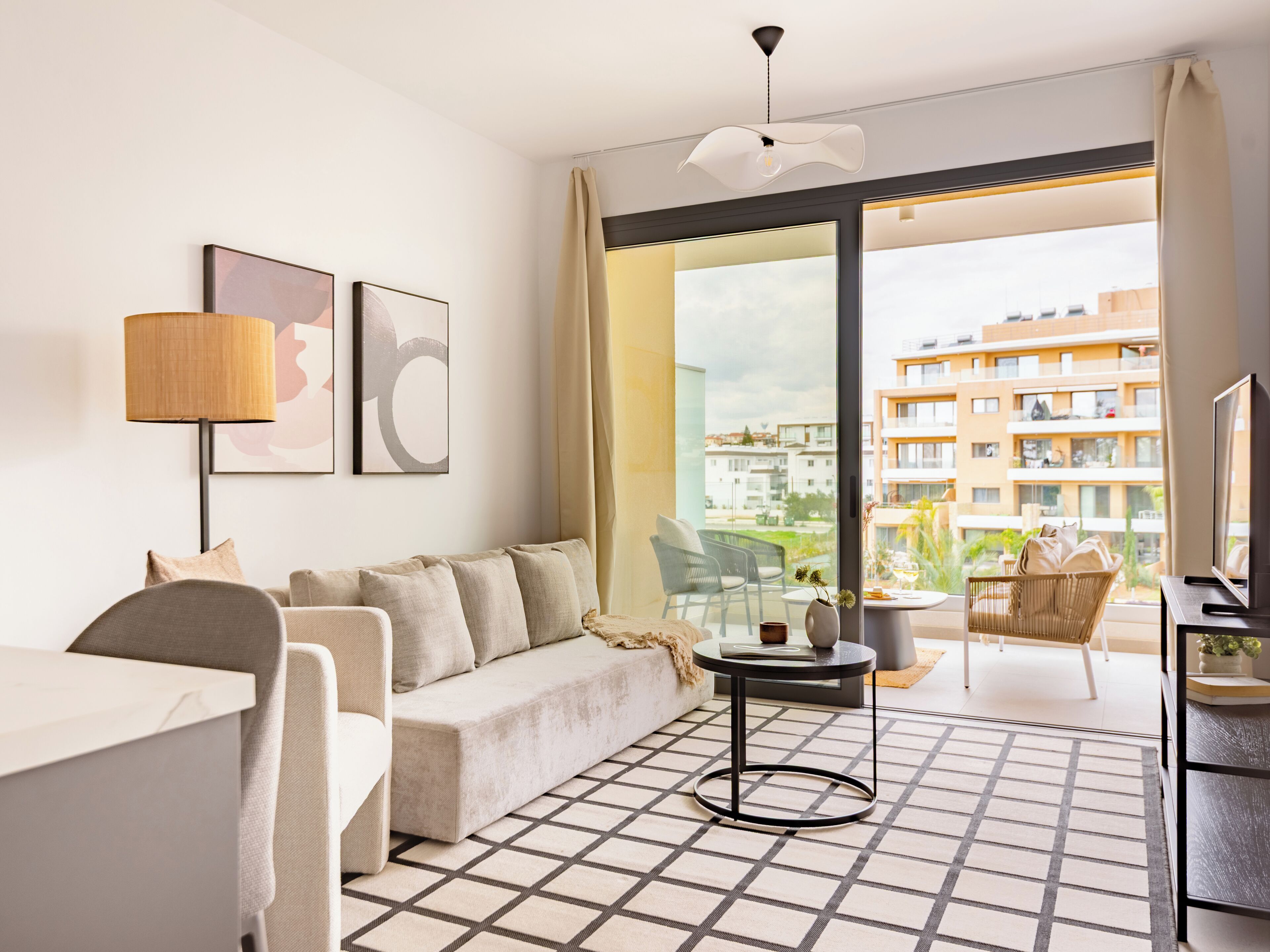 A modern hotel room with a sofa, a small table, a TV, and a balcony with a view of the city.