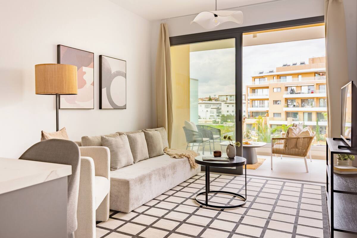 A modern hotel room with a sofa, a small table, a TV, and a balcony with a view of the city.