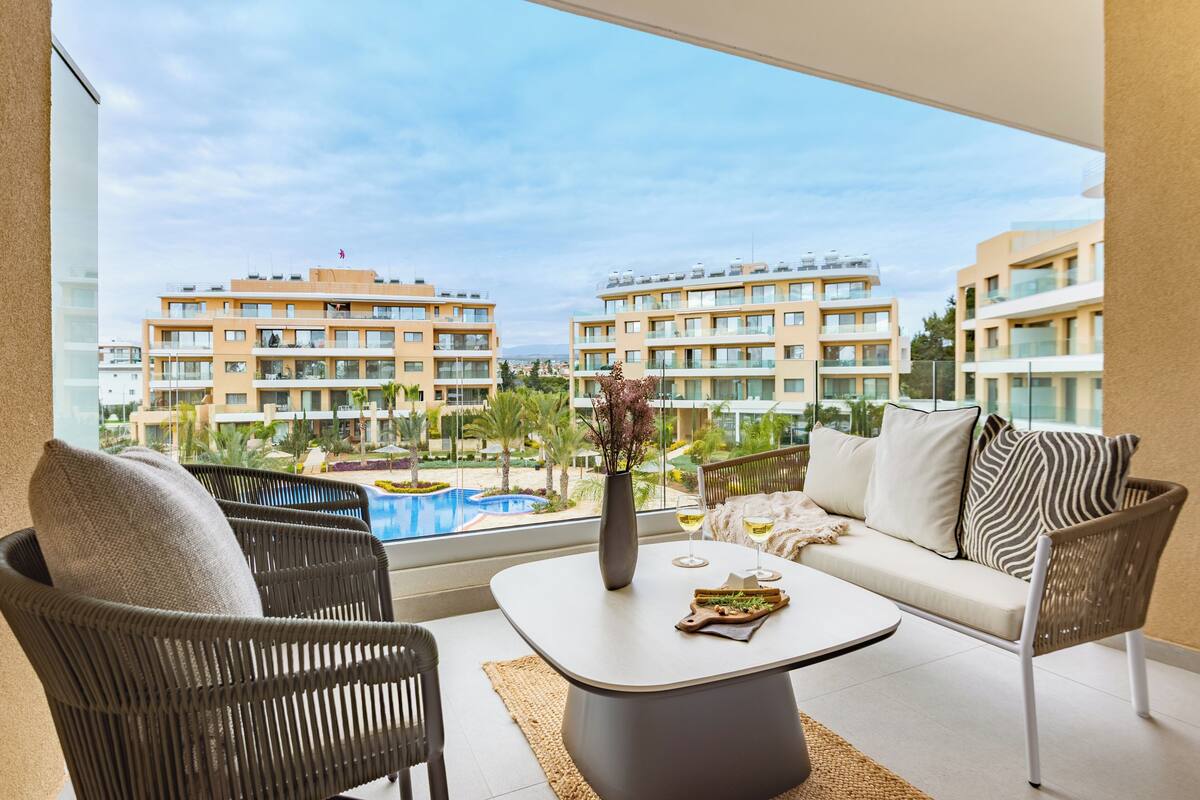 A balcony with wicker chairs, a sofa, and a table with a vase and wine glasses. A pool and buildings are visible in the background.