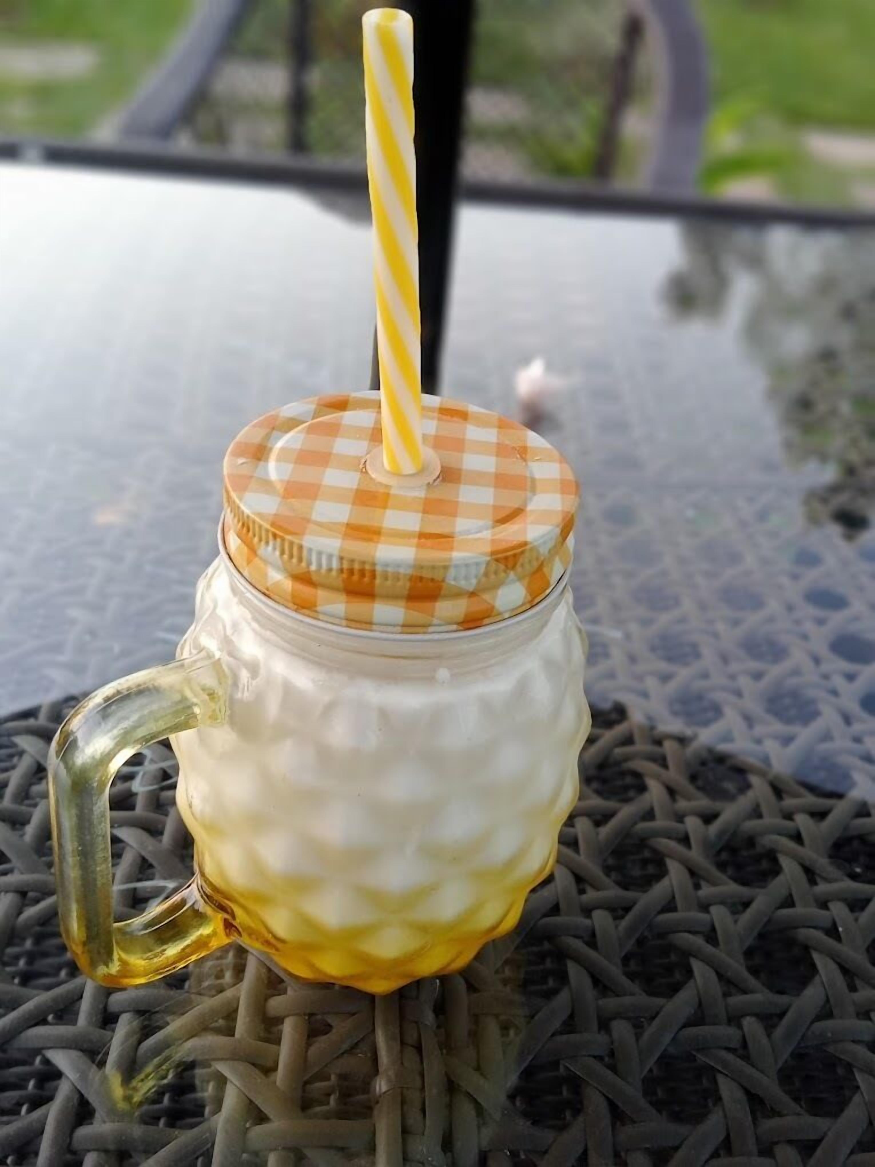 Un frasco de vidrio con tapa a cuadros y una pajita rayada de amarillo, sobre una mesa de metal con superficie trenzada.