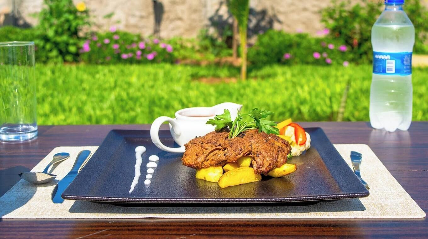 A meal on a black plate with a steak, fries, and a side salad, accompanied by a white mug and a bottle of water on a table with a garden view.
