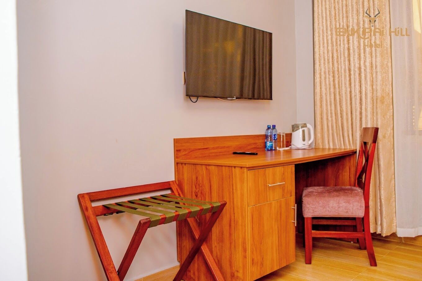 Een compacte kamer met een houten bureau, een stoel, een aan de muur bevestigde televisie, een raam met gordijnen, en een klein tafeltje met een waterkoker en flessen.