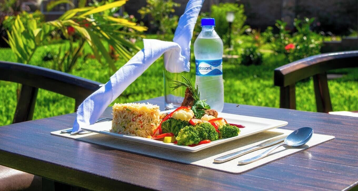 A meal on a table with a bottle of Dasani water, a piece of rice, vegetables, and a piece of meat, with a view of greenery and a building in the background.