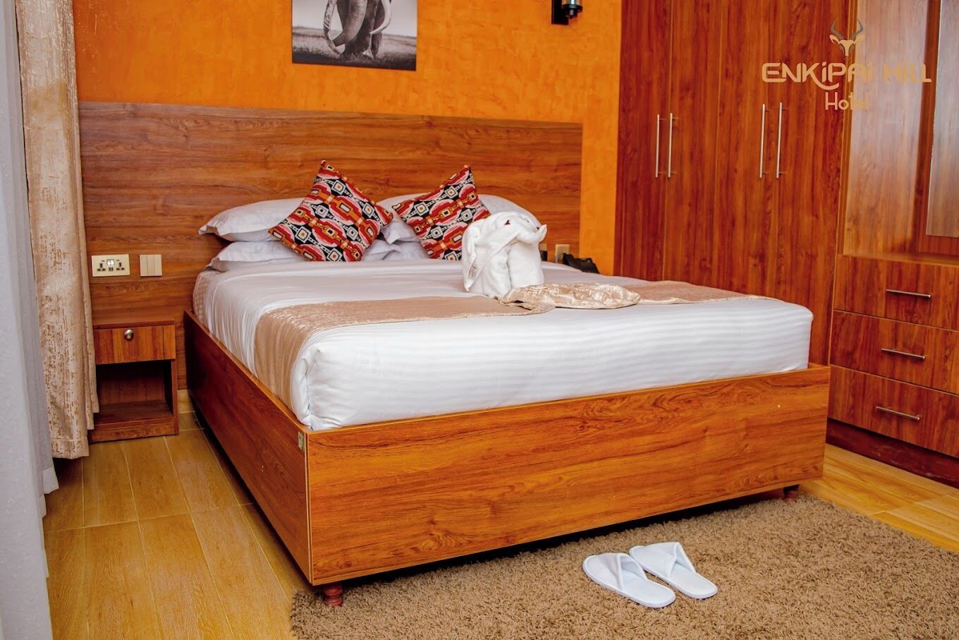 Un lit en bois avec des coussins à motifs, une couverture blanche et une paire de chaussons posés sur le sol.