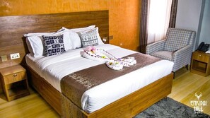 A neatly made bed with a brown throw blanket, white bedding, and decorative towels. There is a wooden nightstand with a lamp, a patterned armchair, and a small table with a phone. - Enkipai Hill Hotel (Narok)