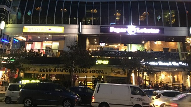 A multi-story building with illuminated windows at night, a modern glass facade, and a ground-level commercial area with signage for a restaurant and a beer ATM. - Urbano Luxury Homestay (George Town)