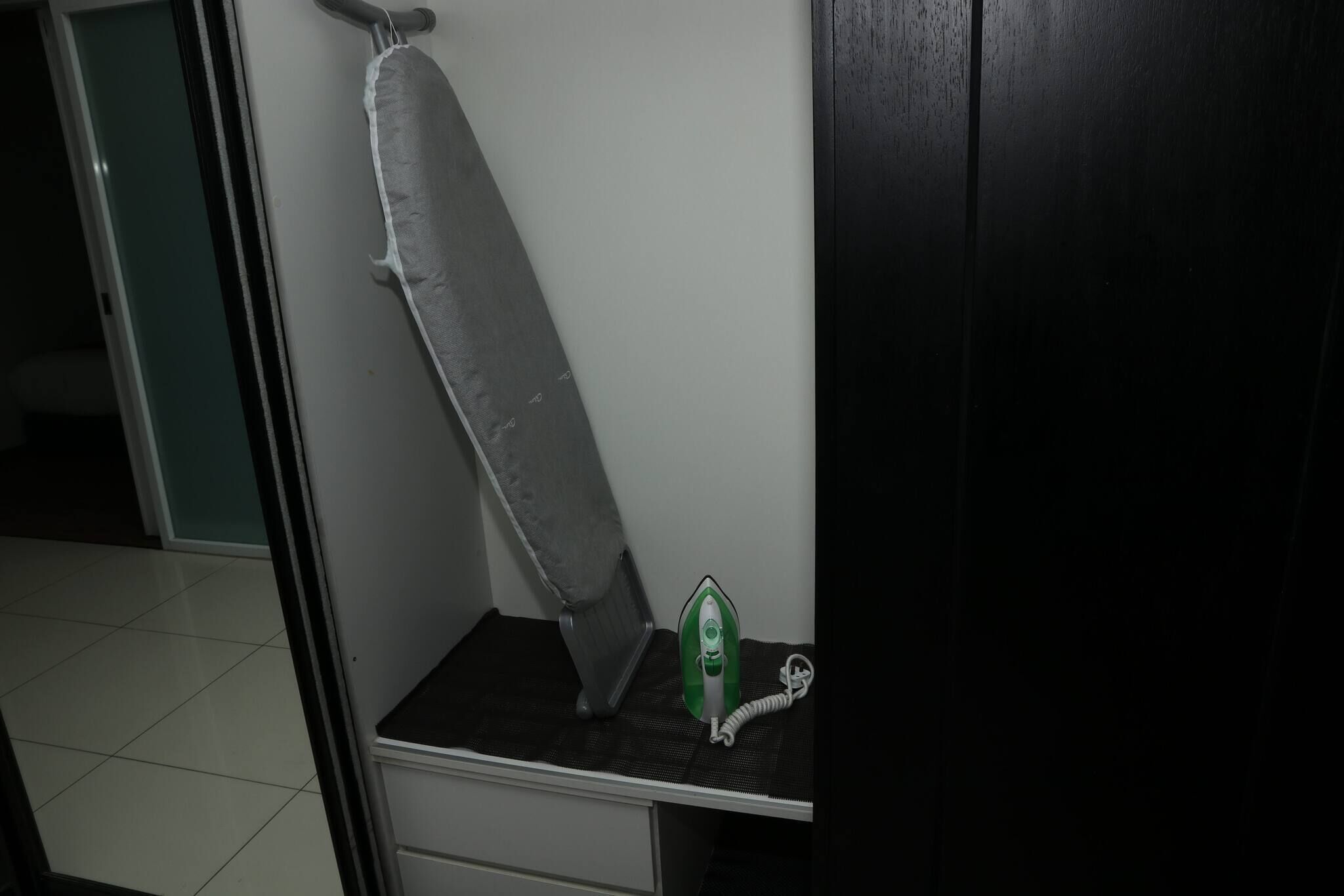 A room with a white ironing board, a green iron, and a white drawer unit.