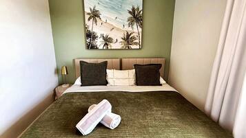 A hotel room with a bed, pillows, a painting of a beach scene, and a rolled towel.