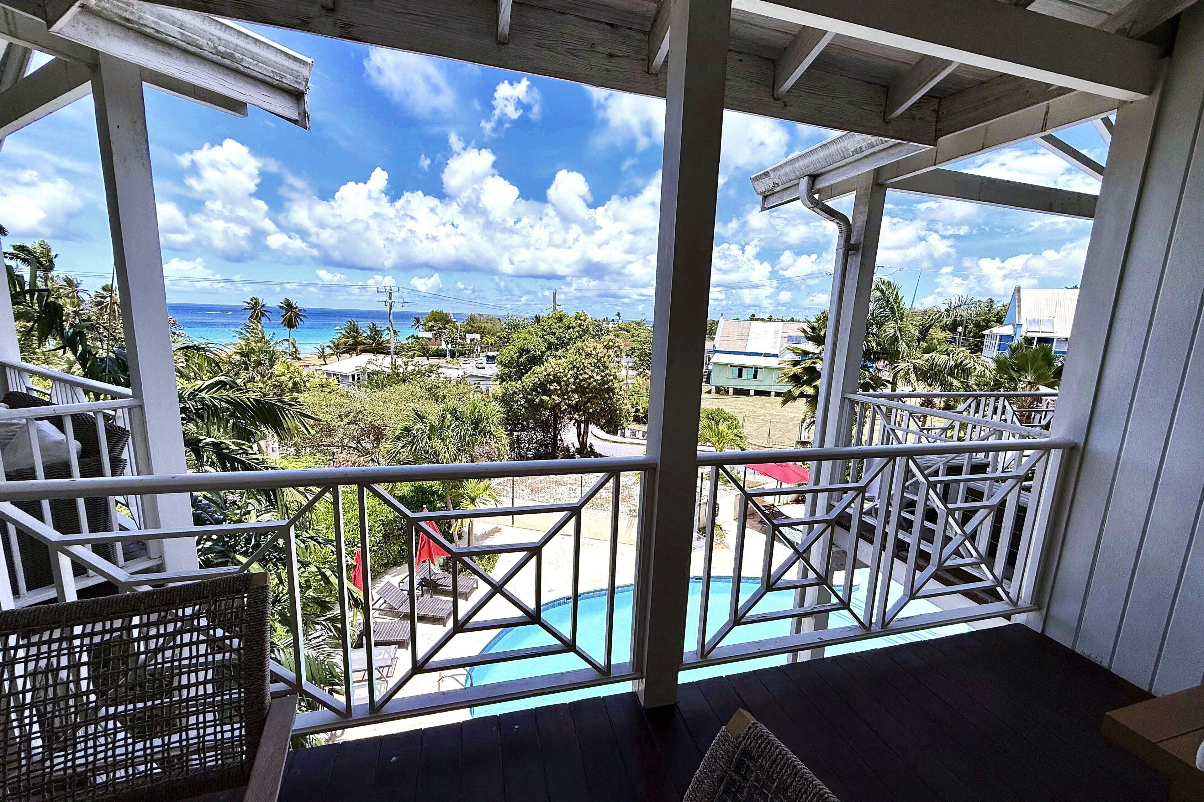 A covered outdoor area with a pool, wooden deck, and a view of the ocean and palm trees.