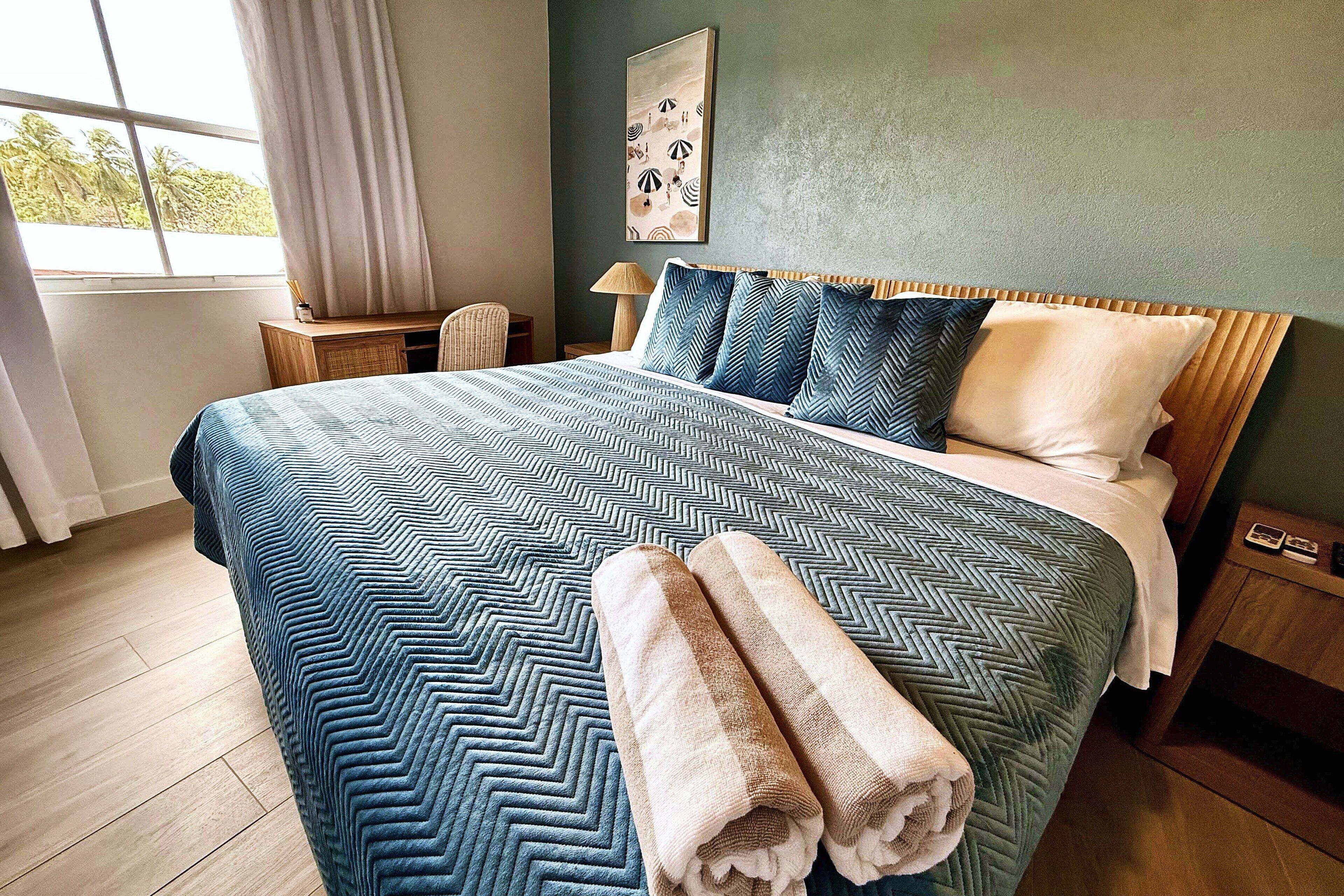 A hotel room with a large bed, a bedside table, a lamp, and a view of the outdoors through a window.
