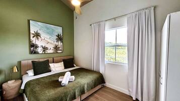 A bedroom with a bed, a painting of a beach scene, a window with curtains, and a ceiling fan.