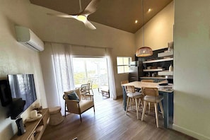 A spacious living area with a ceiling fan, a flat-screen TV, a dining table with chairs, and a kitchen area with a hanging light fixture. - Lantana Sea La Vie - by Zenbreak (Weston)